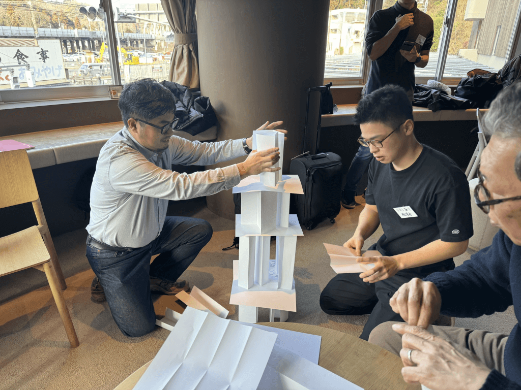 数人に分かれたチームメンバーが白い紙を使ってペーパータワーを集中しながら組み立てています。